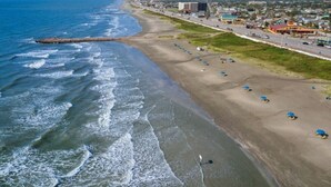 A coastal beach with a long stretch of sand, a breakwater, and a road with buildings and vehicles. - Cozy Family Home, Prime Location Near Downtown (Galveston)