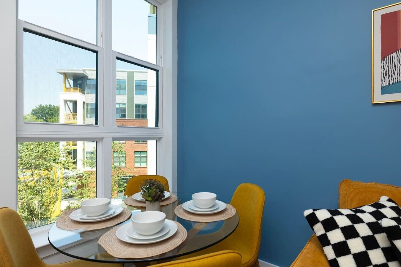 Comedor con una mesa dispuesta para cuatro, sillas amarillas y vista a edificios a través de amplios ventanales.