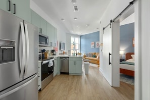 A modern kitchen with stainless steel appliances, a central island, and a view into a living area with a sofa and a bed.