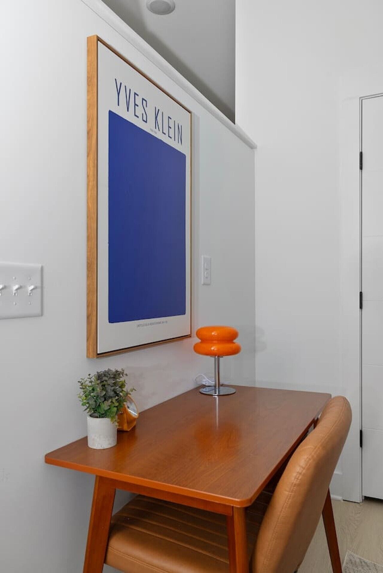 Ein Essbereich mit einem Holztisch, einem einzelnen orangefarbenen Stuhl, einem kleinen Topfpflanzen und einem gerahmten Poster an der Wand.