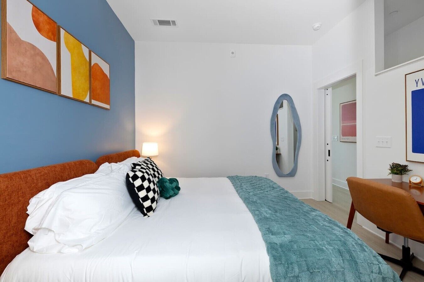 A hotel room with a bed, a desk, a chair, a mirror, and artwork on the walls.
