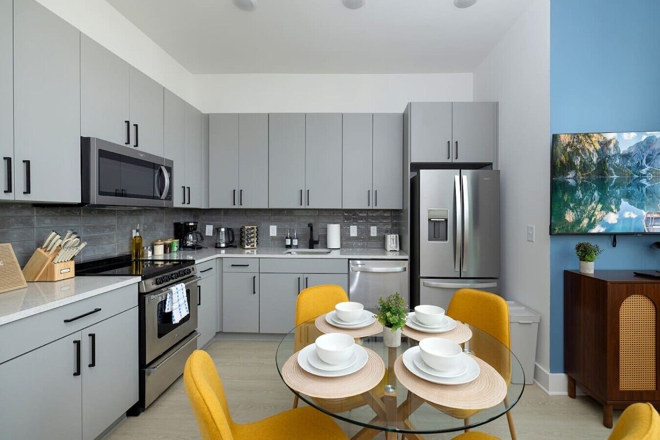 A modern kitchen with grey cabinets, stainless steel appliances, and a dining area with yellow chairs and a glass table.