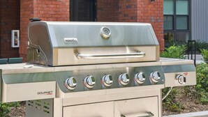 Uma churrasqueira a gás Gourmet Pro da NEXGRILL com acabamento em aço inoxidável, localizada ao ar livre em frente a um edifício de tijolos.