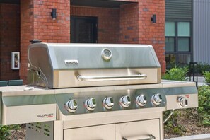 A NEXGRILL Gourmet Pro gas grill with a stainless steel finish, located outdoors in front of a brick building. - 1BR King w/ Gym + Courtyard w/ Grill Station + Free Parking, NoDa Art District (Charlotte)