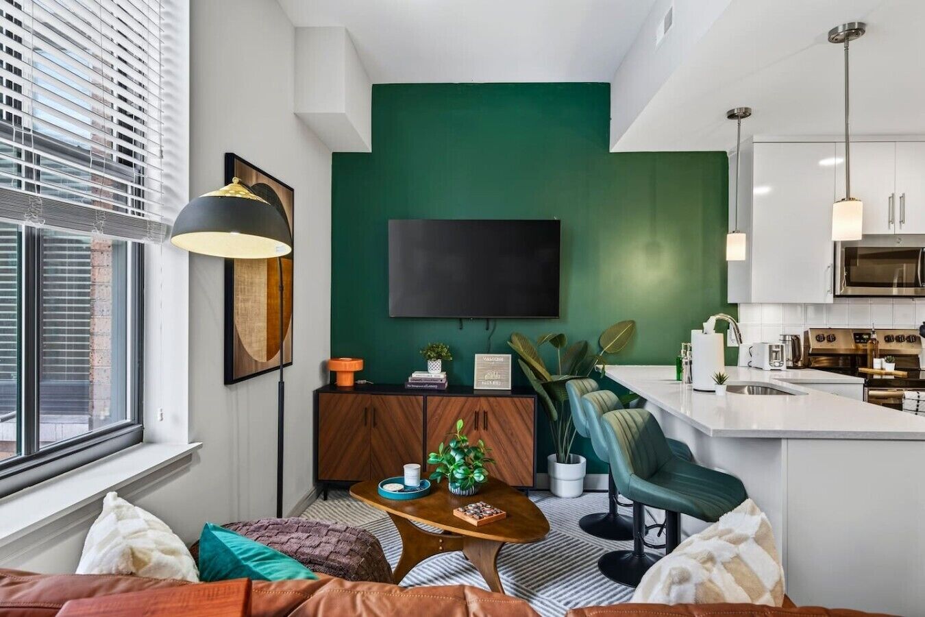 A modern living room with a green accent wall, a flat-screen TV, a wooden cabinet, and a kitchen area with a microwave and stove.