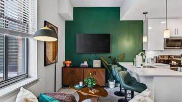 A modern living room with a green accent wall, a flat-screen TV, a wooden cabinet, and a kitchen area with a microwave and stove.
