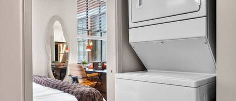 A compact hotel room with a bed, a small kitchenette with a stove, and a view of the city through the window.