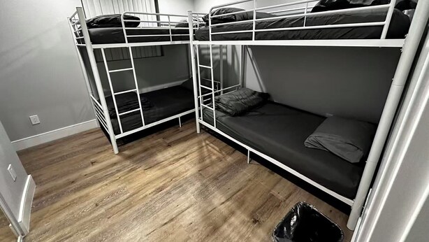 Une chambre avec un lit superposé, un plancher en bois, une fenêtre avec des stores et une poubelle.