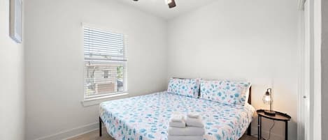 A bedroom with a bed, a ceiling fan, a window with blinds, and a bedside table with a lamp.