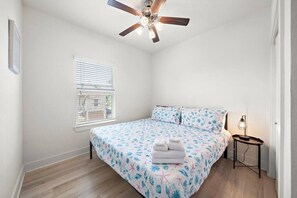 A bedroom with a bed, a ceiling fan, a window with blinds, and a bedside table with a lamp. - Cozy Family Home with Game Room, Near Downtown (Galveston)