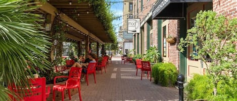 Un café en plein air avec des chaises et des tables rouges, un bâtiment en briques sur lequel on peut lire l’inscription « THÉ ET ESPRESSO », et de la verdure.
