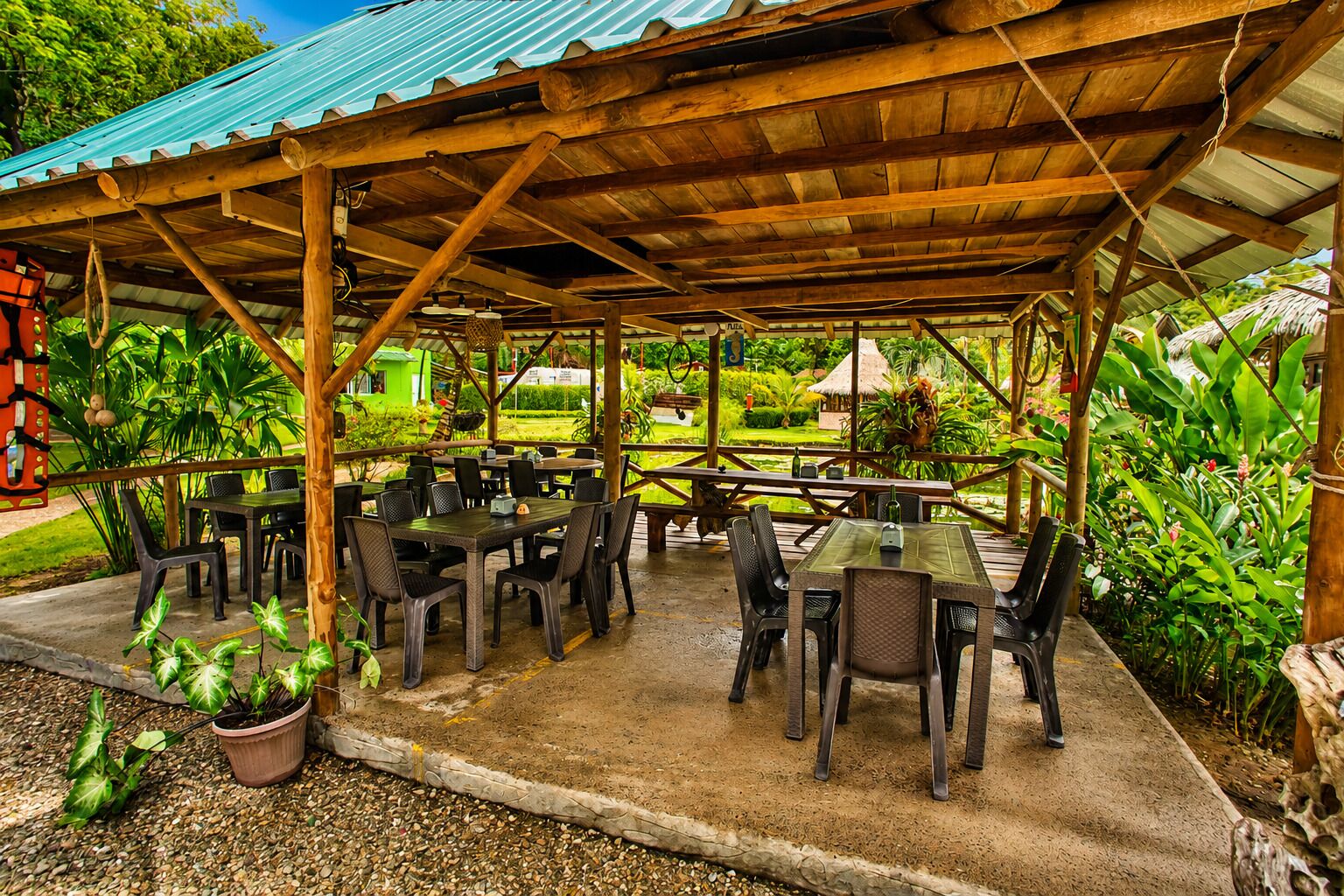 An outdoor covered dining area with tables and chairs, surrounded by greenery and a thatched-roof structure in the background.