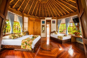 Una habitación con dos camas, suelo de madera, techo de paja y ventanas grandes con cortinas.