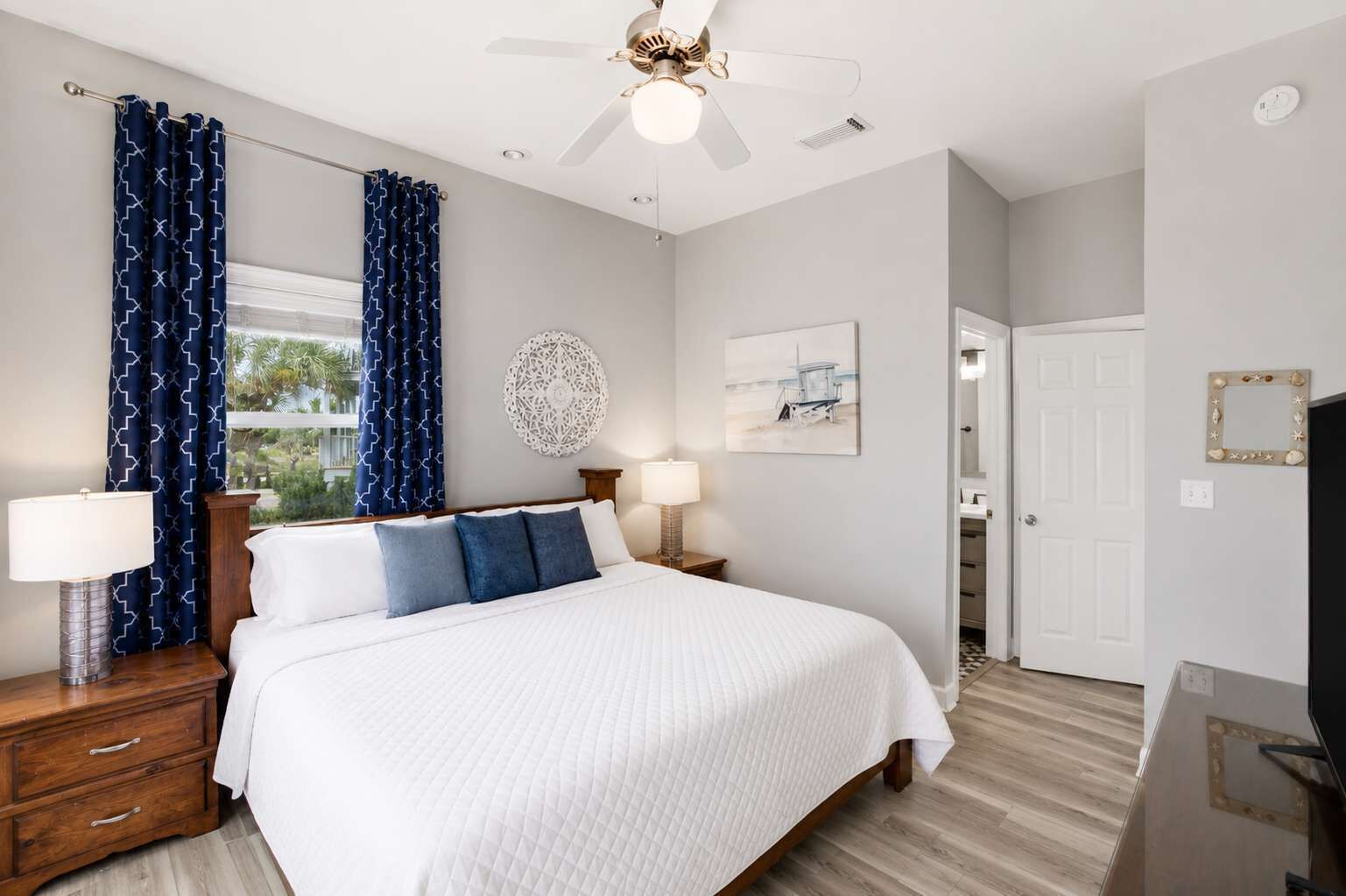 A hotel room with a bed, two bedside tables, a ceiling fan, a window with curtains, a wall clock, and a door leading to a bathroom.