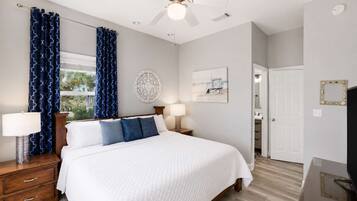 A hotel room with a bed, two bedside tables, a ceiling fan, a window with curtains, a wall clock, and a door leading to a bathroom.