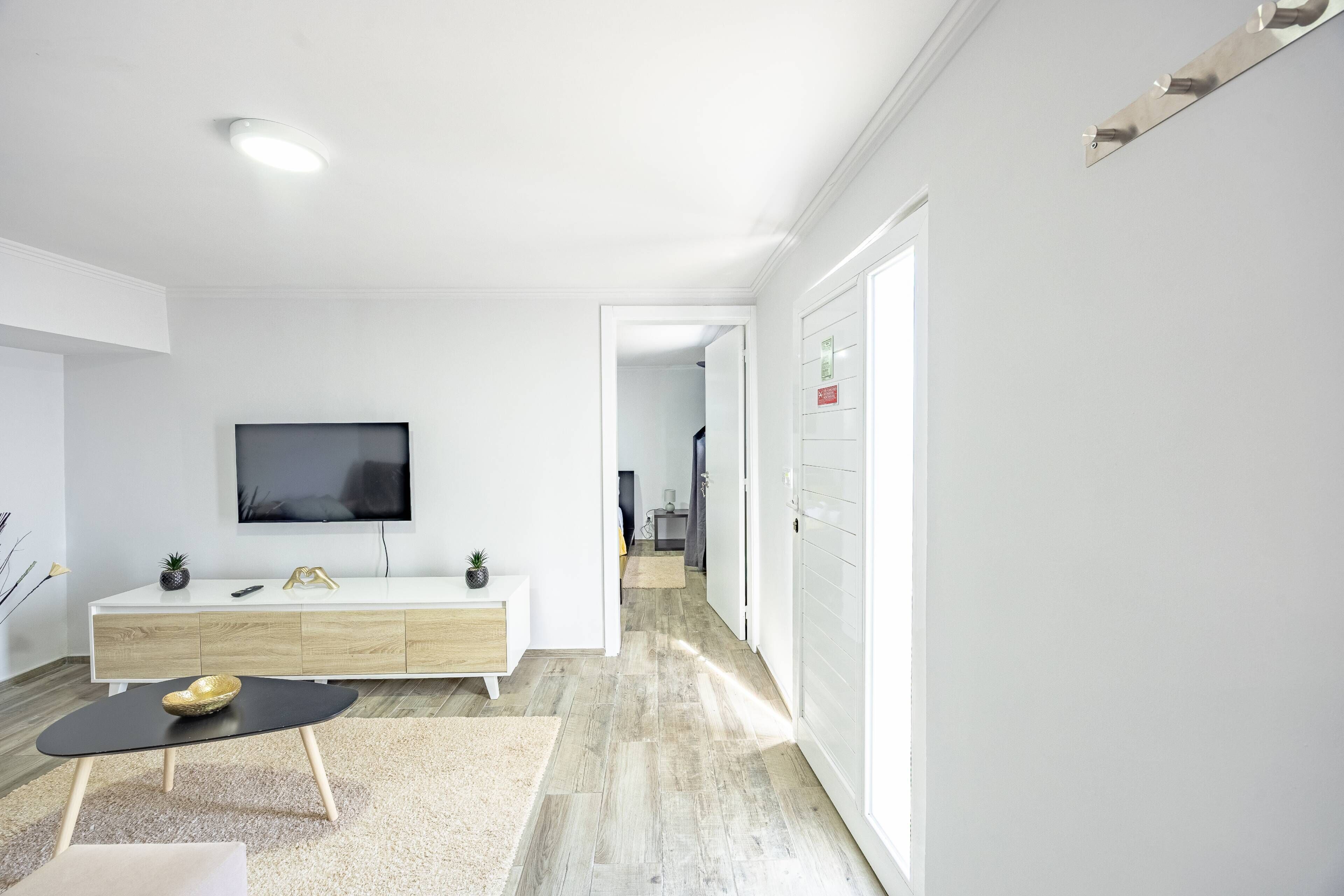 Een moderne, goed verlichte woonkamer met een flatscreen televisie, een witte console tafel, een zwarte salontafel en een houten vloer.