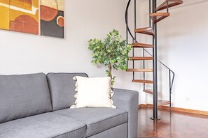Ein graues Sofa mit einem weißen Kissen, ein Blumentopf und eine Wendeltreppe vor einer weißen Wand.