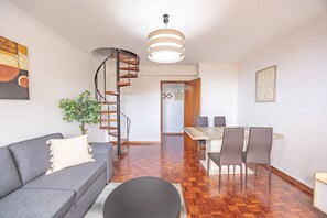 Ein modernes Wohnzimmer mit einem grauen Sofa, einem schwarzen runden Tisch, einer Holzspirale und einem Essbereich mit Holztisch und Stühlen.