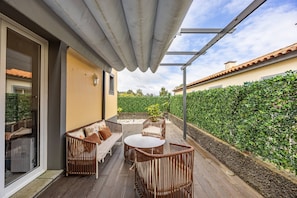 Eine Terrasse im Freien mit Korbmöbeln, einer Pergola und Blick auf einen grünen Heckenstreifen.