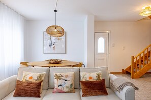 A living room with a round wooden table, a sofa with decorative cushions, a hanging woven light fixture, and a door with a window. - Cozy Haven Santana by YOUR KEY (Santana)