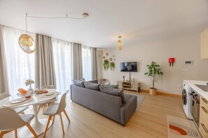 A modern living room with a grey sofa, dining table, and chairs. There is a flat-screen TV on a stand, a washing machine, and a potted plant. - Fénix Horizon Suite by YOUR KEY (Santa Cruz)