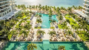 Een resort met een centraal gelegen zwembad, omringd door ligbedden en palmbomen, met uitzicht op een strand en de oceaan.
