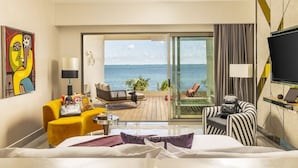 A hotel room with a bed, a yellow armchair, a striped armchair, a TV, and a balcony with a view of the sea.