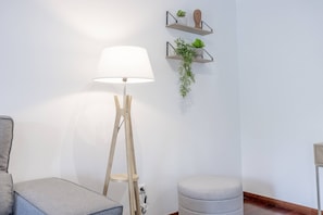 A room with a standing floor lamp, a grey sofa, a round ottoman, and two wall-mounted shelves with plants and decorative items. - Funchal Haven by YOUR KEY (Funchal)