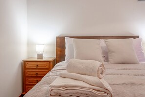 A neatly made bed with a folded blanket and pillows, a wooden nightstand with a lamp, and a wall-mounted light. - L&P Countryside by YOUR KEY (Santana)
