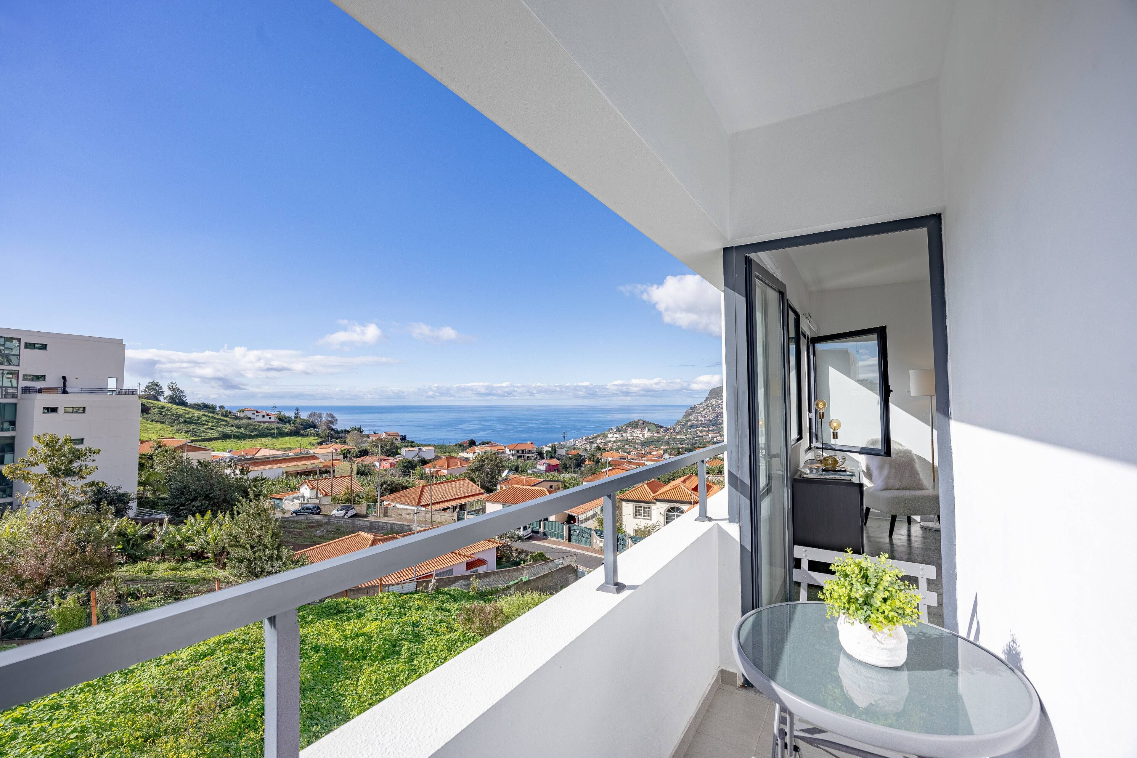 A balcony with a glass table, a chair, and a view of a coastal town.