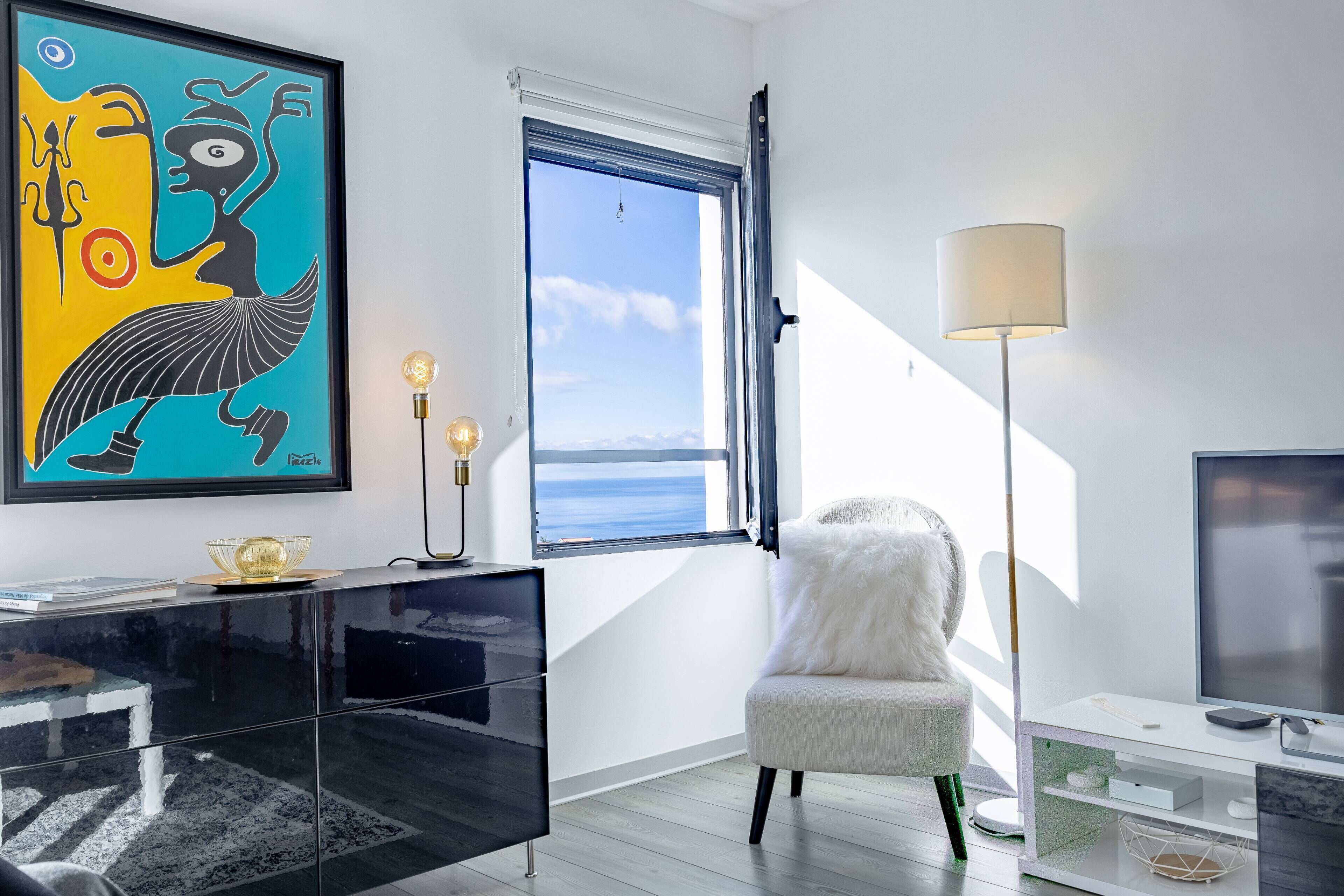 Una sala de estar moderna con un ventanal grande, un cuadro abstracto enmarcado, una silla blanca, una lámpara de pie y un televisor de pantalla plana.