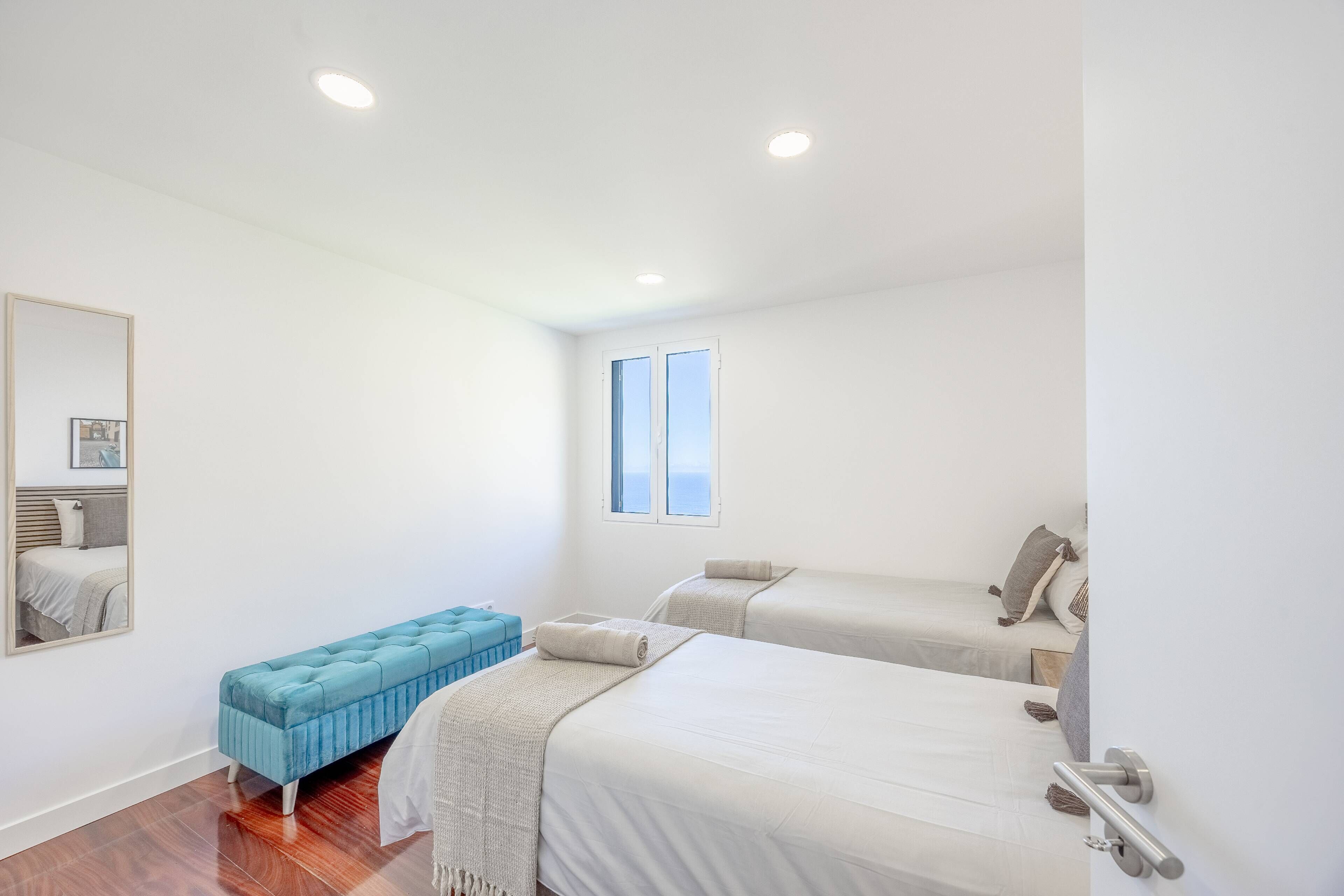 A bedroom with two beds, a bench, a window, and a mirror.