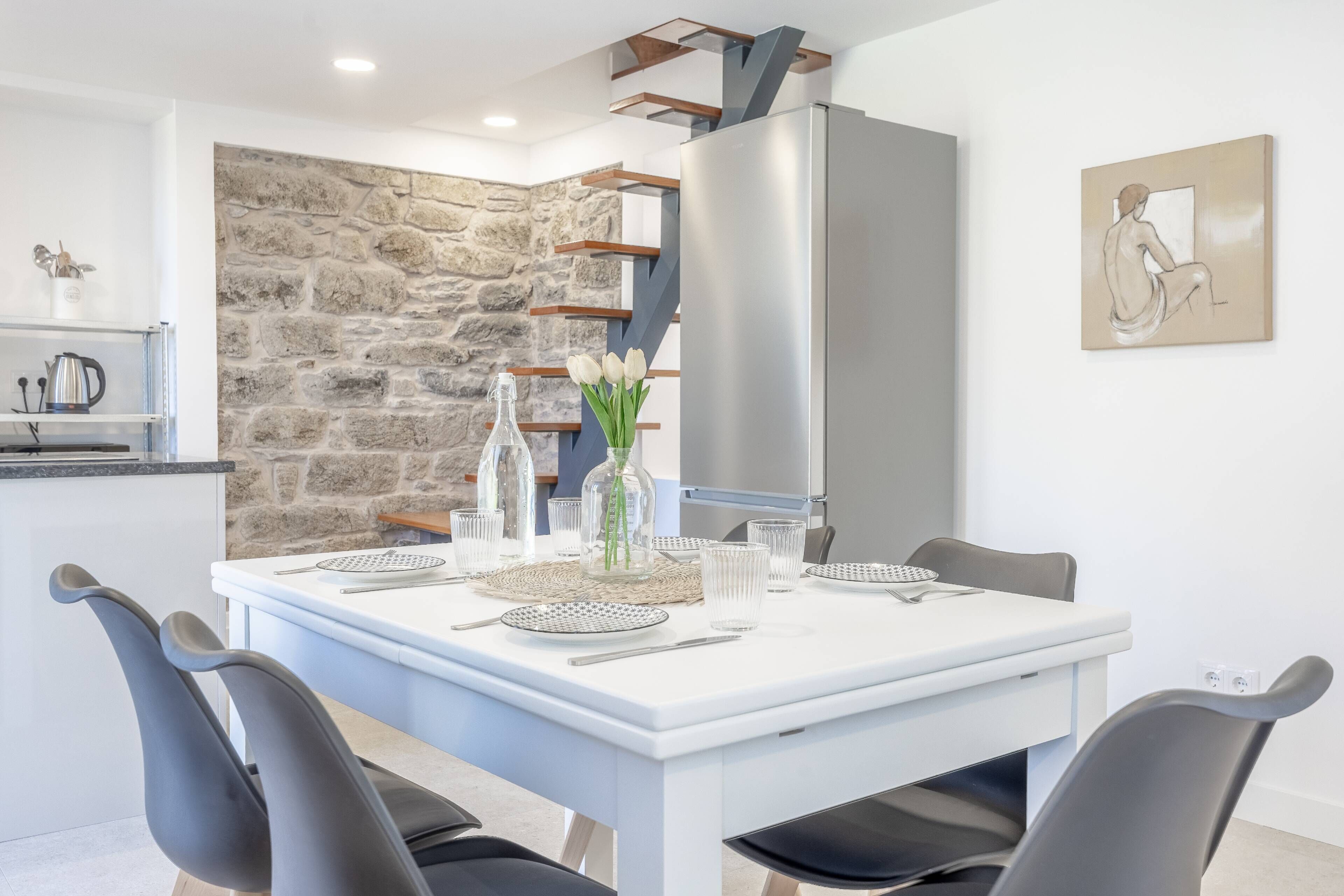 A dining area with a white table set for four, surrounded by black chairs. A stone wall and a modern kitchen with a stainless steel refrigerator are visible in the background.
