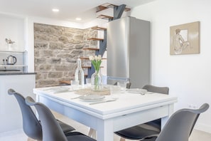 Comedor con una mesa blanca dispuesta para cuatro personas, rodeada de sillas negras. Se aprecia una pared de piedra y una cocina moderna con un refrigerador de acero inoxidable al fondo.