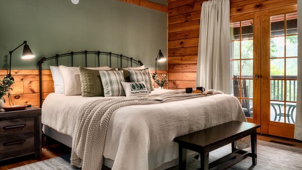 A wooden cabin-style bedroom with a bed, bedside lamps, a bench, and a window with curtains. - VIEWS! / Hot Tub / Nintendo Switch / Games! (Sevierville)