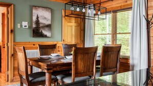 A dining area with a wooden table and chairs, a painting on the wall, and a window with curtains. - VIEWS! / Hot Tub / Nintendo Switch / Games! (Sevierville)