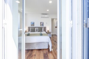 Ein modernes Schlafzimmer mit einem großen Bett, einem Nachttisch und einem Beistelltisch. Ein Fenster mit Jalousien, eine Wand-Klimaanlage und zwei gerahmte Bilder an der Wand sind vorhanden.