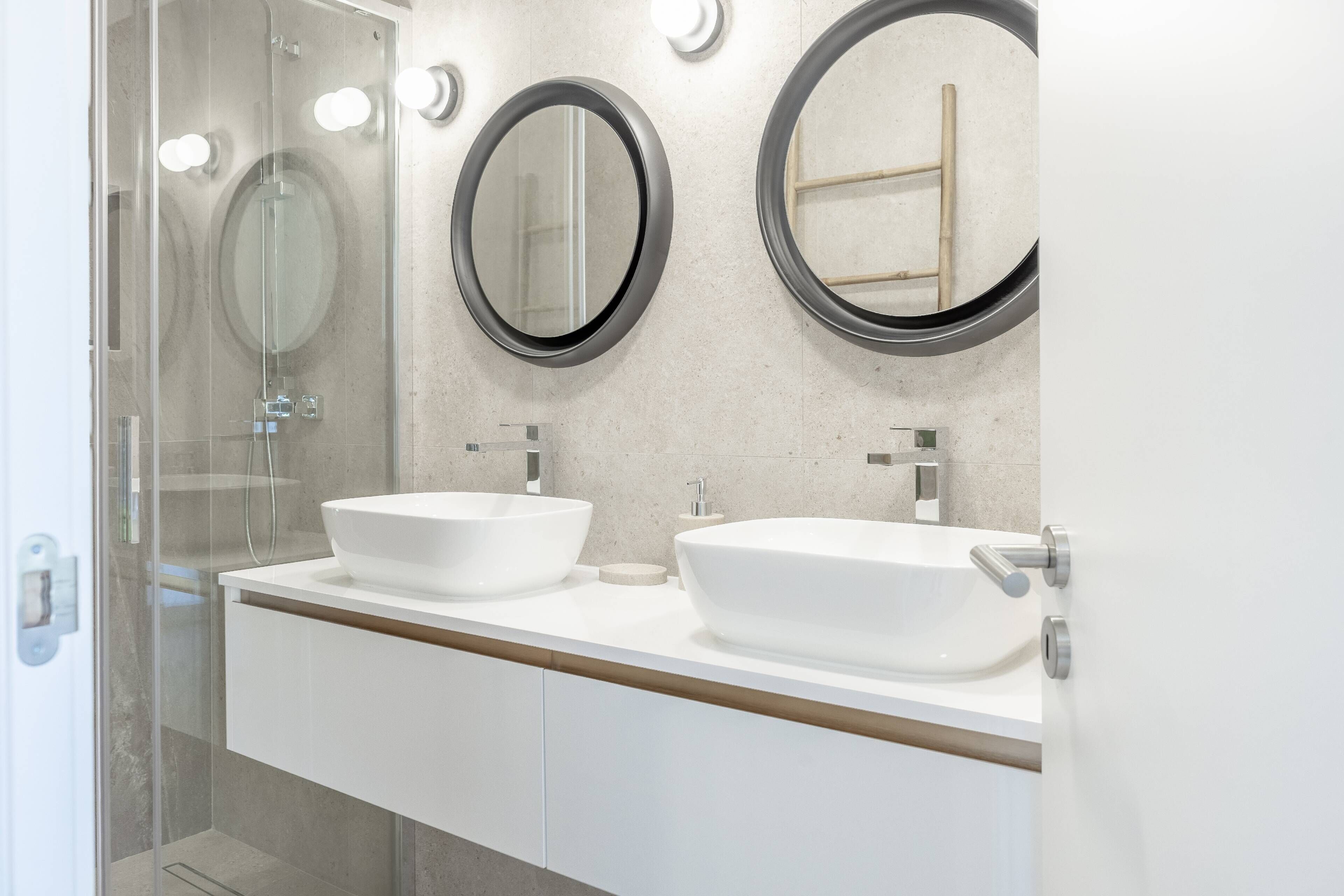 Une salle de bain avec deux lavabos blancs, une douche en verre et deux miroirs ronds à cadre noir.