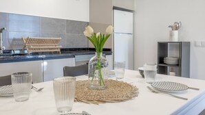 A modern kitchen with a dining table set for four, featuring clear glassware, a woven placemat, and a vase with white flowers. - Maison Des Souvenir B by YOUR KEY (Ribeira Brava)