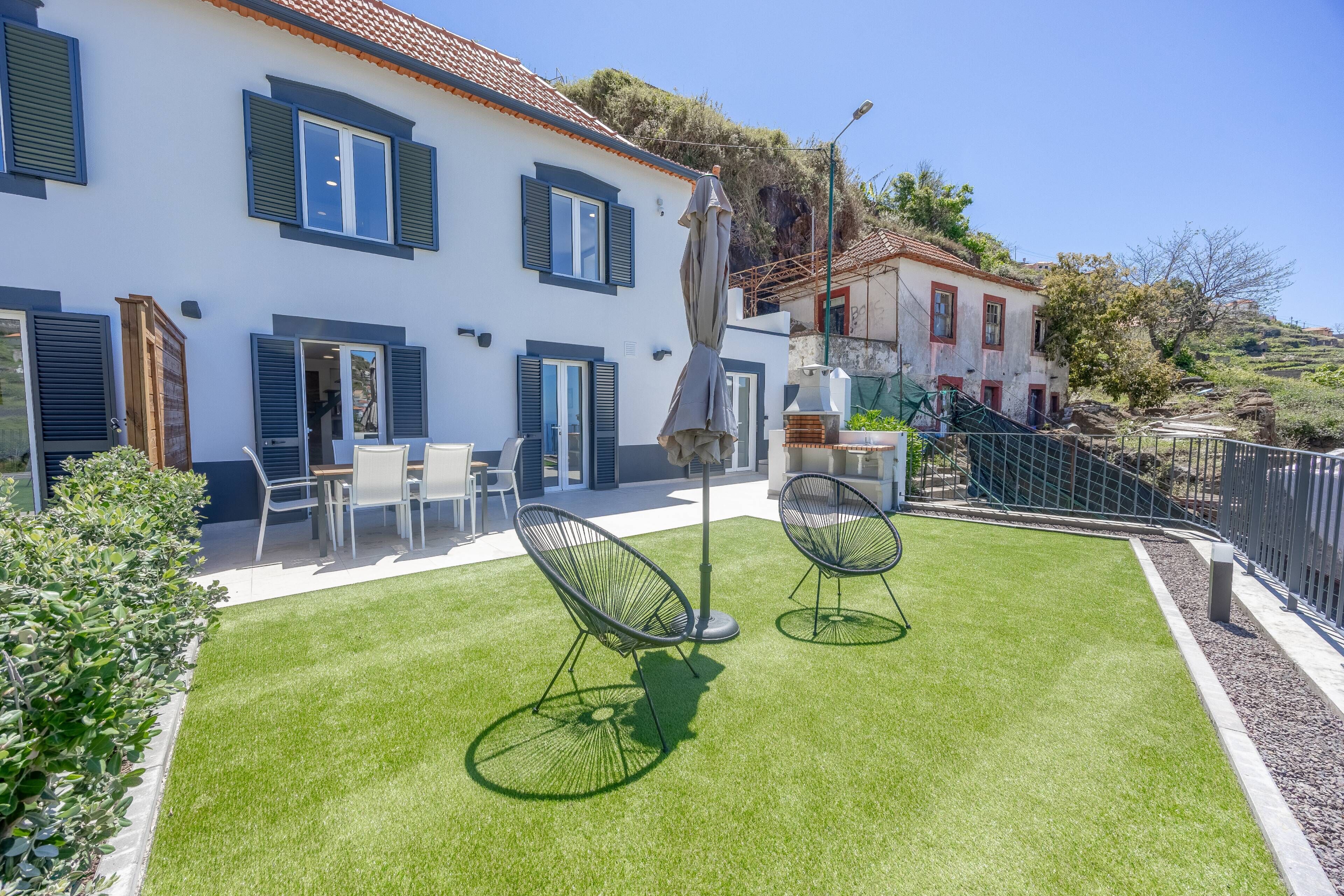 Propiedad residencial con césped cuidado, zona de estar exterior y un edificio blanco con contraventanas azules.