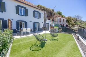 Propiedad residencial con césped cuidado, zona de estar exterior y un edificio blanco con contraventanas azules.