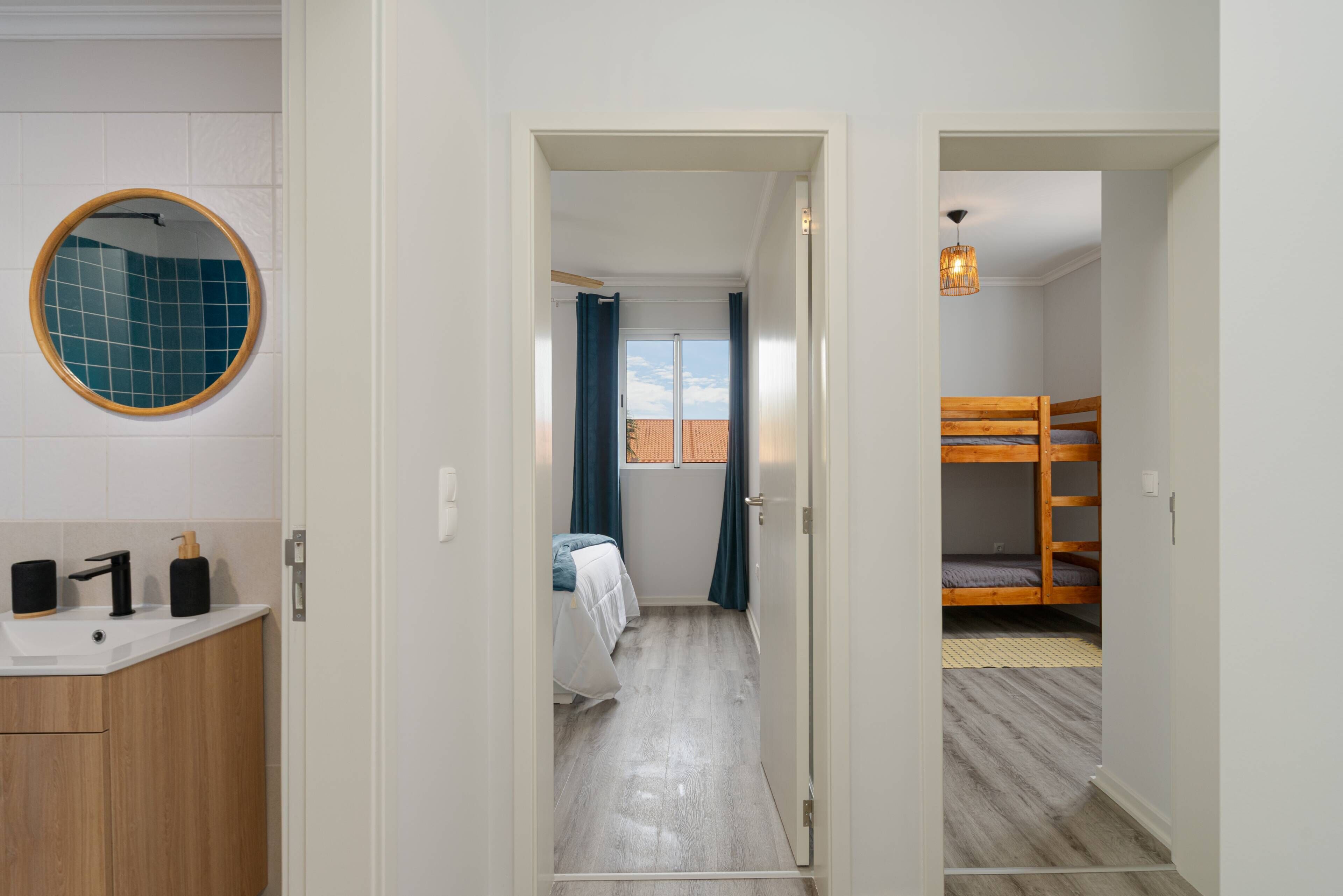 Een badkamer met een wastafel, spiegel en toiletartikelen. Achter een open deur is een slaapkamer zichtbaar met een bed en een raam.