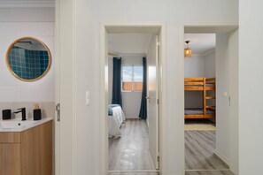 A bathroom with a sink, mirror, and toiletries. A bedroom with a bed and a window is visible through an open door. - MJ - North Escape by YOUR KEY (Porto Moniz)