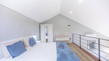 A modern bedroom with a sloped ceiling, a large bed with a blue blanket, a crib, and a balcony with a railing.