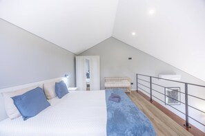 A modern bedroom with a sloped ceiling, a large bed with a blue blanket, a crib, and a balcony with a railing.