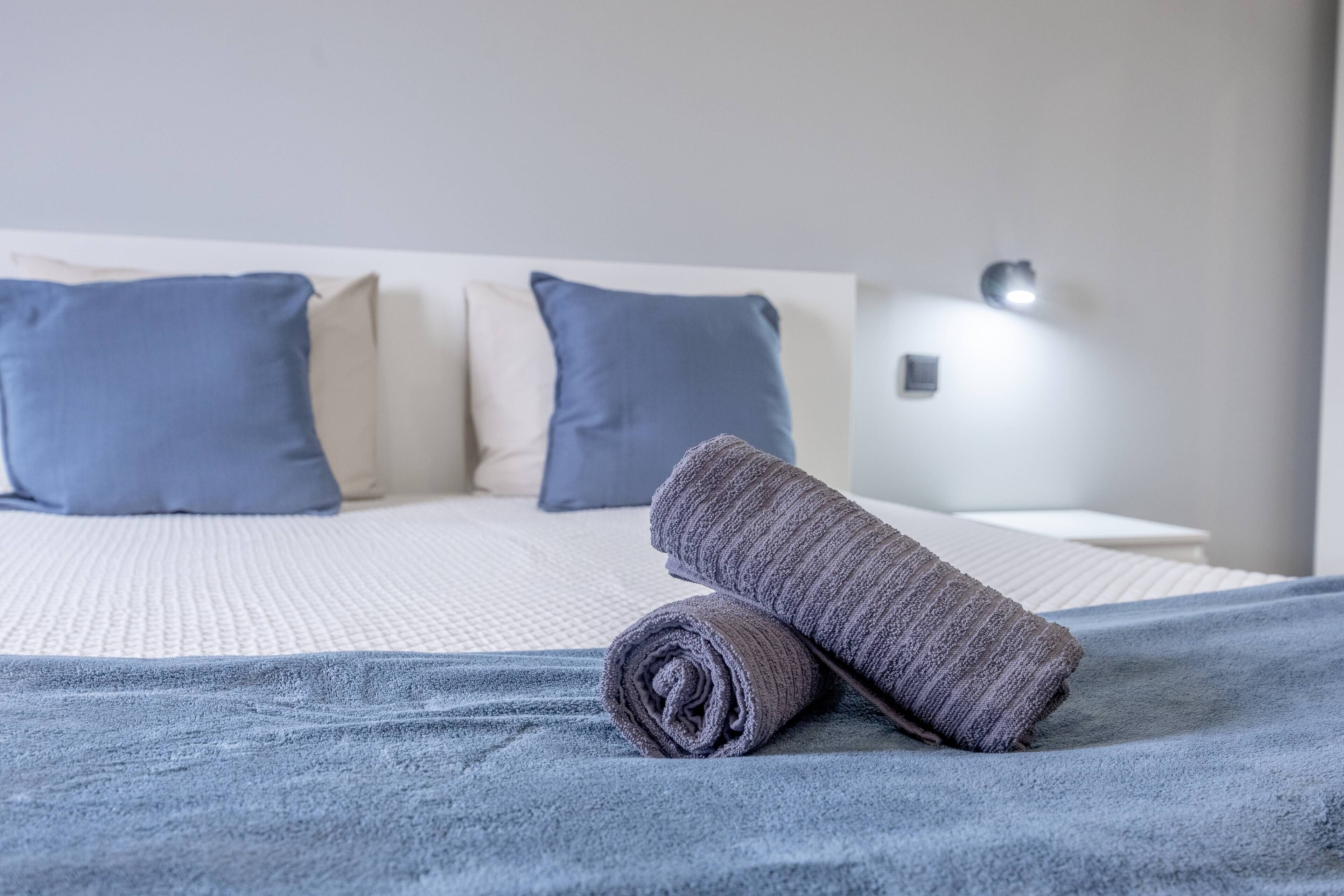 Ein ordentlich bezogenes Bett mit einem aufgerollten Handtuch darauf, einem weißen Kopfteil und einer Nachttischlampe.