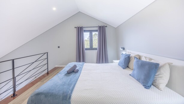 Ein Schlafzimmer mit schrägen Wänden, einem Bett mit blauer Decke und einem Fenster mit Vorhängen.