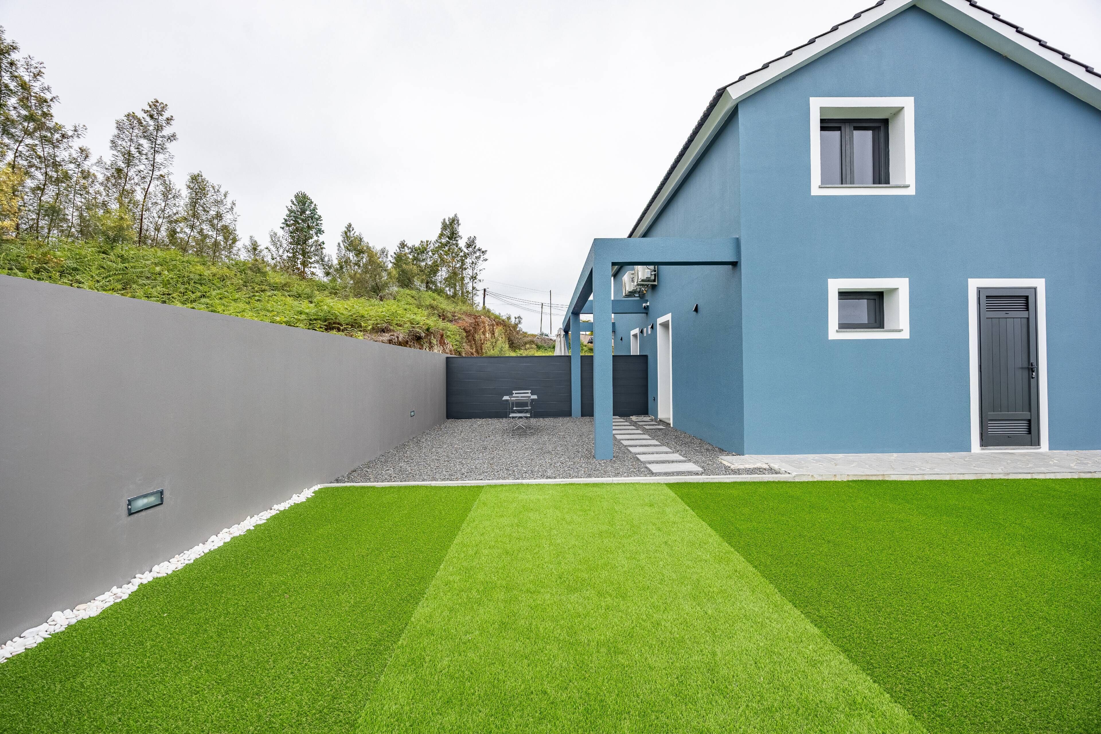 Ein modernes blaues Haus mit grauer Wand, einer kleinen Terrasse mit Kunstrasen und einem Kiesbereich.