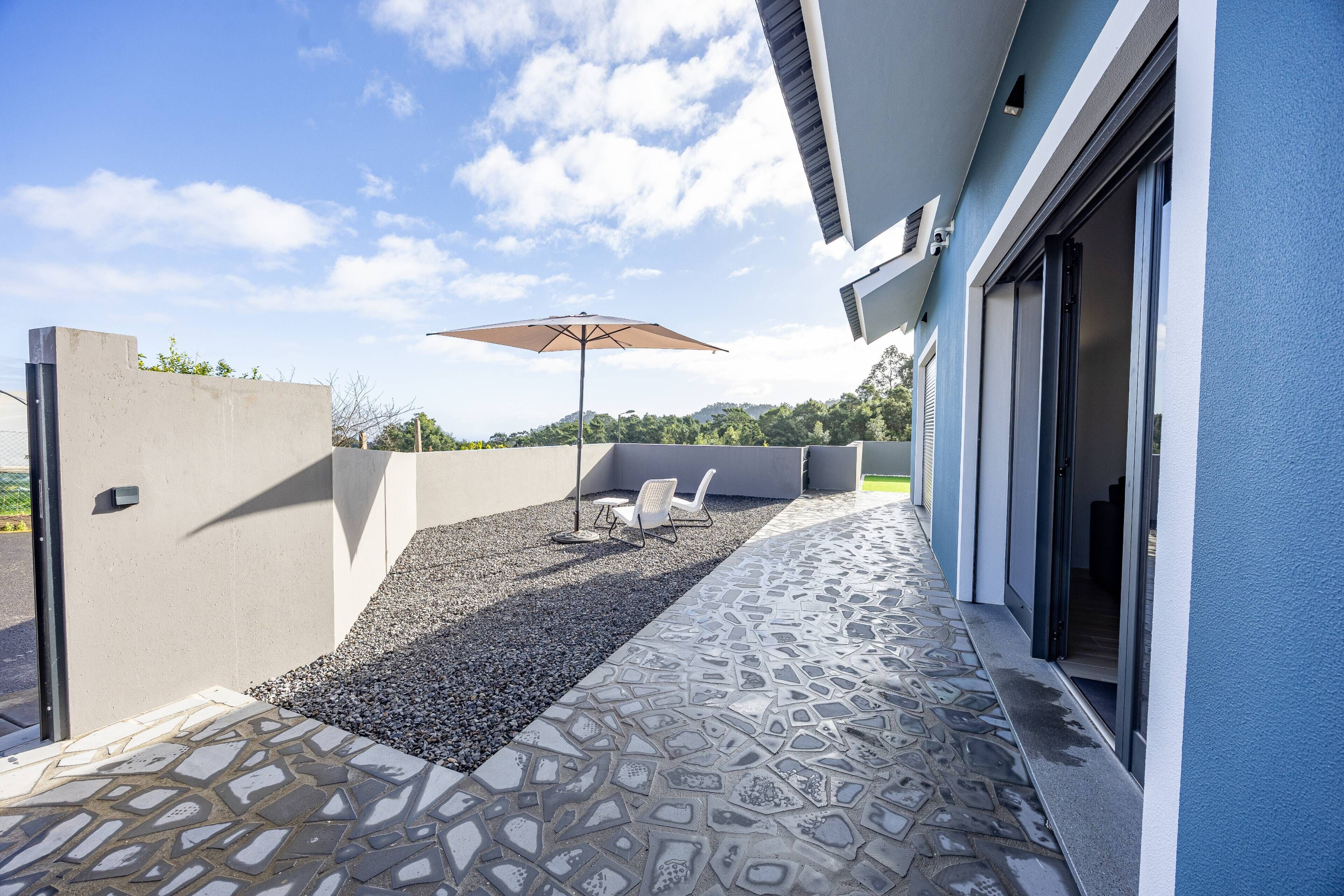 A patio area with a patterned stone floor, a concrete wall, a seating area with a chair and an umbrella, and a building entrance with a sliding door.