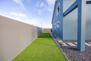 Un sendero estrecho con césped artificial que lleva a un edificio con fachada azul y una pared gris a un lado.